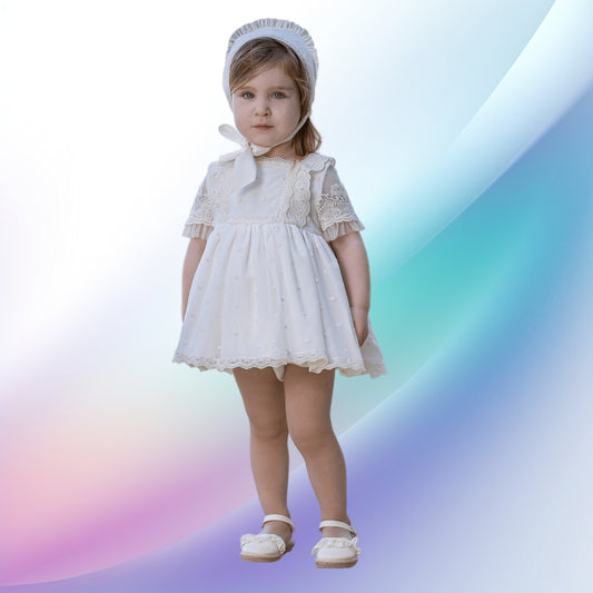 An image of an elegant off-white baby and toddler ceremony dress, perfect for a baptism or church event. The European-style handcrafted dress features a box neckline, delicate lace details, tulle sleeves, and a multilayered skirt with embroidered tulle and lace. A matching bonnet and off-white knickers complete this charming ensemble.
